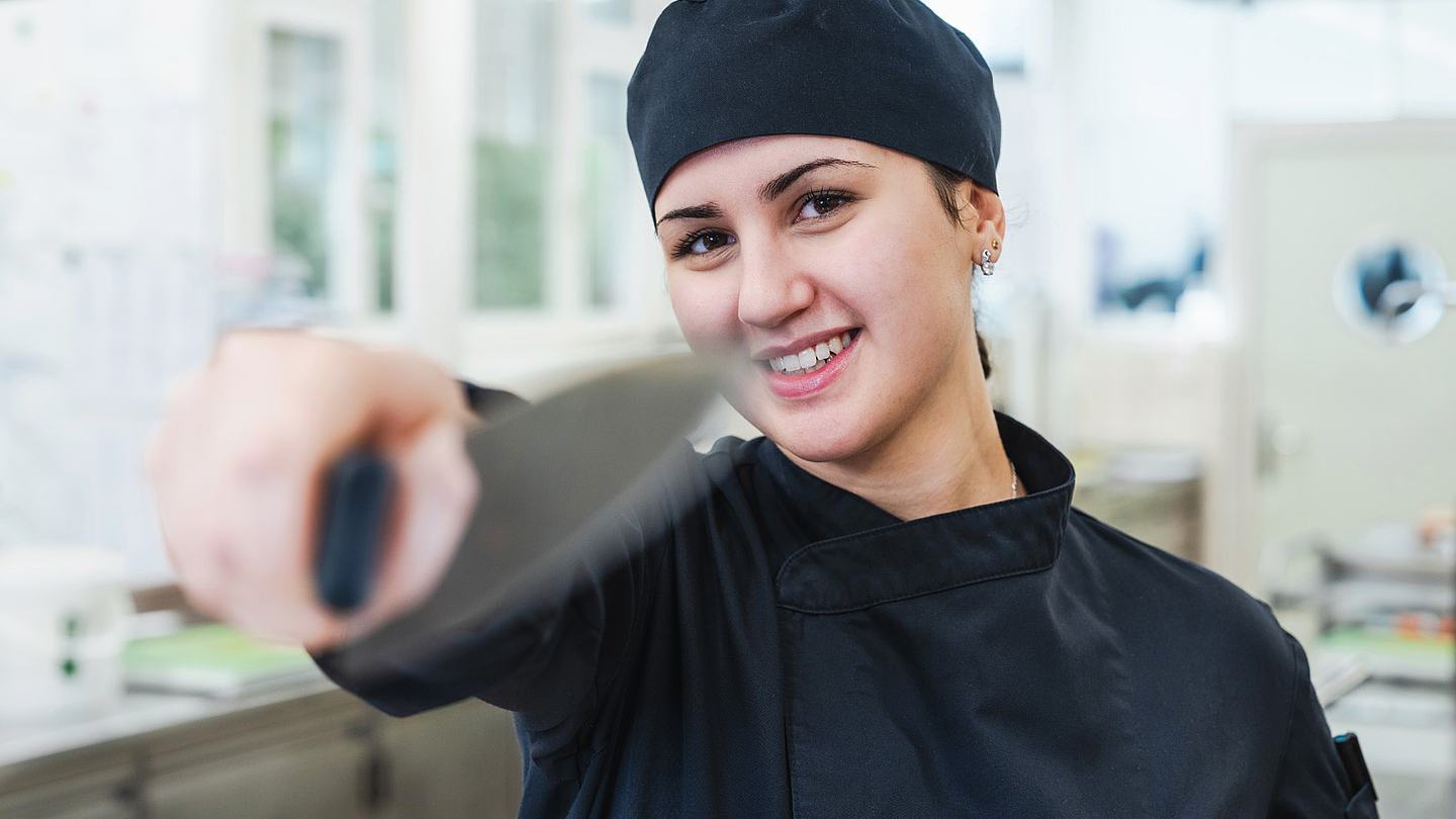 Rodana Mahmoud, Lernende Köchin in der Klinik Meissenberg, steht in der Küche und hält ein Messer in die Kamera.